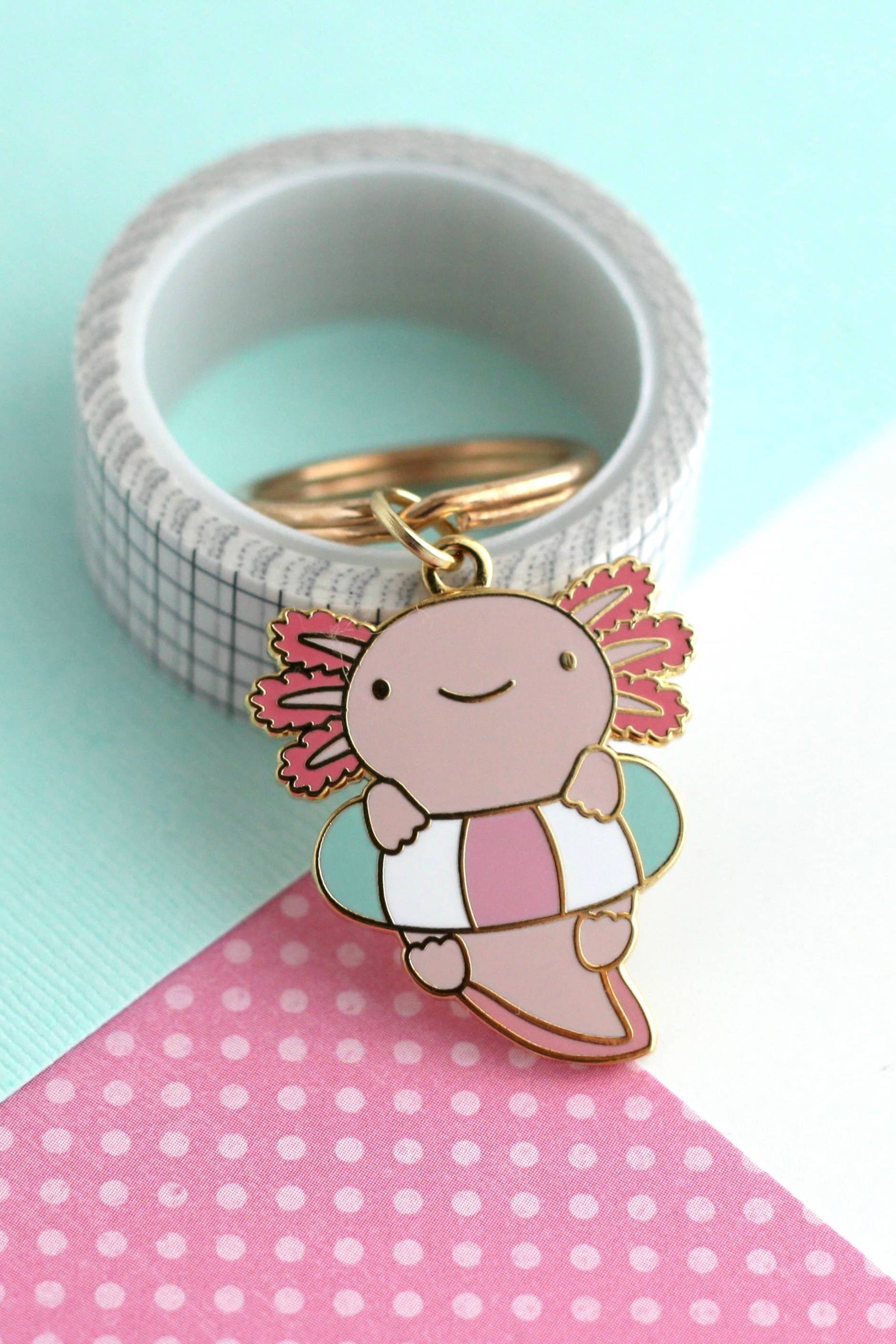 Porte-clés Axolotl Flottant en émail dur doré posé sur fond turquoise et rose à pois, design kawaii pastel.