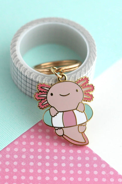 Porte-clés Axolotl Flottant en émail dur doré posé sur fond turquoise et rose à pois, design kawaii pastel.