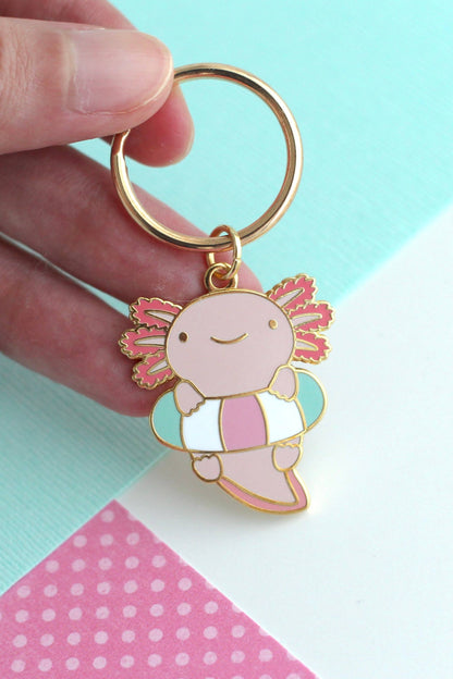 Porte-clés Axolotl Flottant en métal doré, tenu entre les doigts sur fond pastel turquoise et rose.
