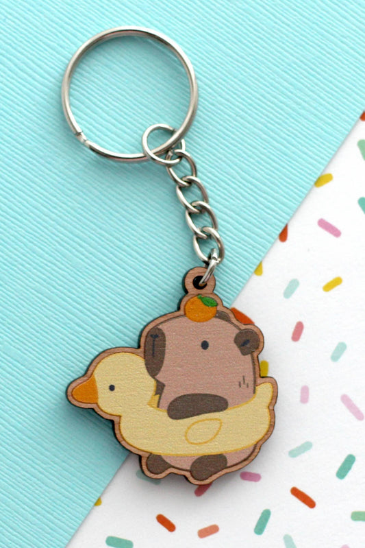 Porte-clés en bois représentant un capybara avec une bouée canard et une orange sur la tête, posé sur fond pastel turquoise et blanc.