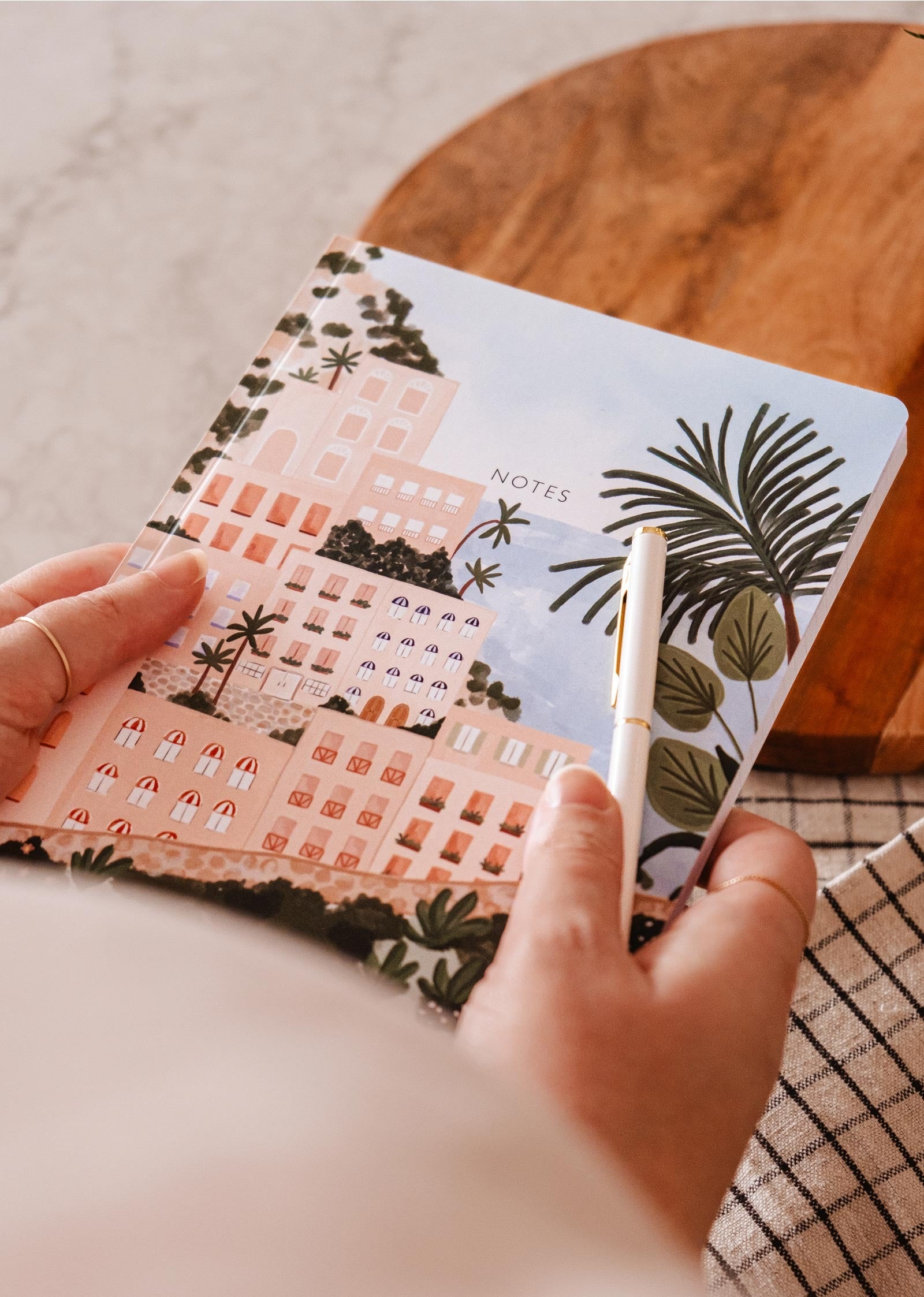 La couverture du carnet de notes est un beau dessin coloré représentant Positano, un ville côtière italienne. Ce sont des grandes maisons roses avec plein de fenêtre qui surplombe la met. ville côtière italienne.Sur la photo, on peut voir tenir le carnet et un stylo dans ses mains. 