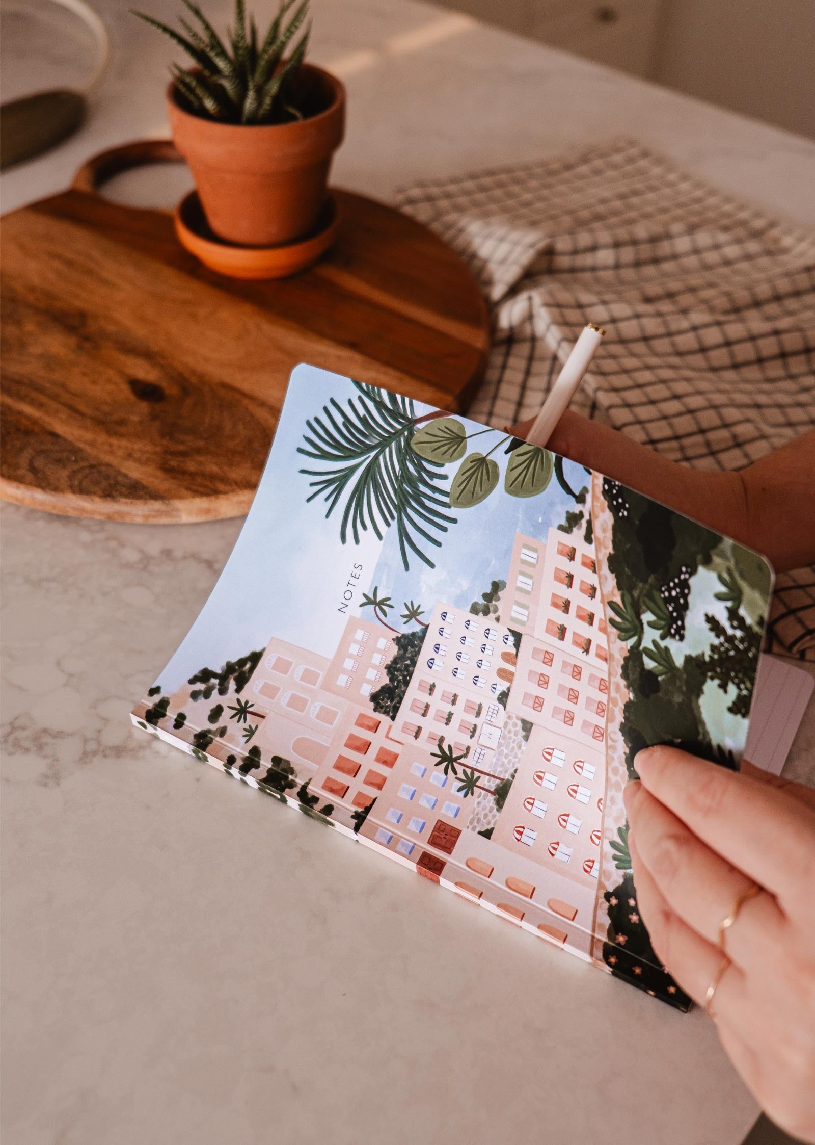 La couverture du carnet de notes est un beau dessin coloré représentant Positano, un ville côtière italienne. Ce sont des grandes maisons roses avec plein de fenêtre qui surplombe la met. ville côtière italienne.Sur la photo, on peut voir la main de quelqu'un qui tient le carnet ouvert. Cette personne est installé à une table en marbre blanc.