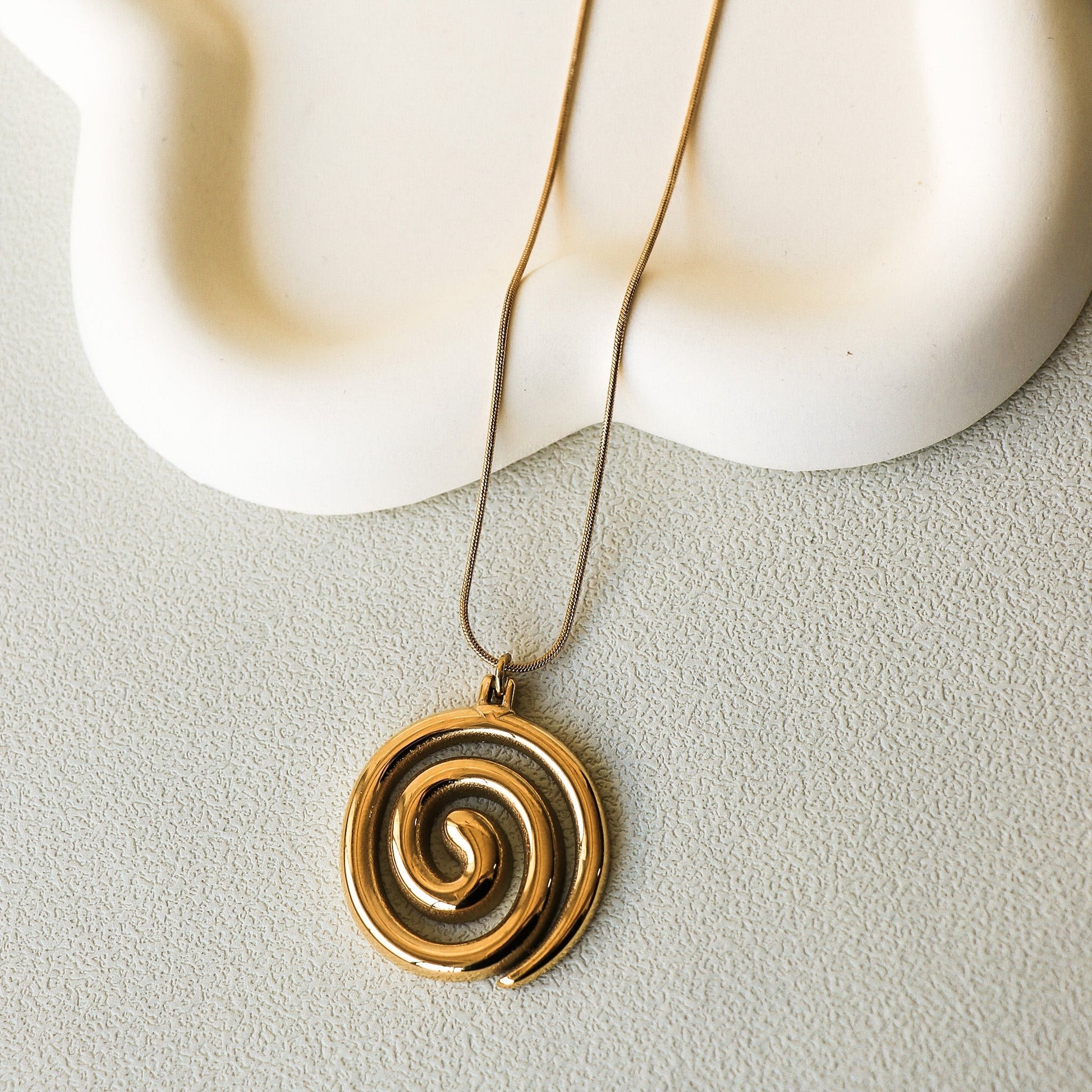 Fine chaîne en acier inoxydable plaqué or avec un pendentif en forme de spirale. Le collier est déposé sur une assiette en céramique blanche en forme d'étoile.