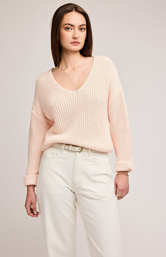 Pull en tricot point demi-cardigan avec un col V et de couleur rose pastel. Le modèle porte un pantalon blanc et une ceinture en cuir aussi blanche. L'arrière-plan est un fond gris.