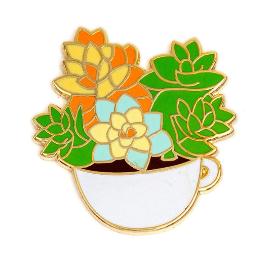 Épinglette en or avec émail coloré en forme de petite tasse blanche contenant des petites succulentes vertes, bleues, jaunes et oranges. de fleurs. L’arrière-plan est un fond blanc.