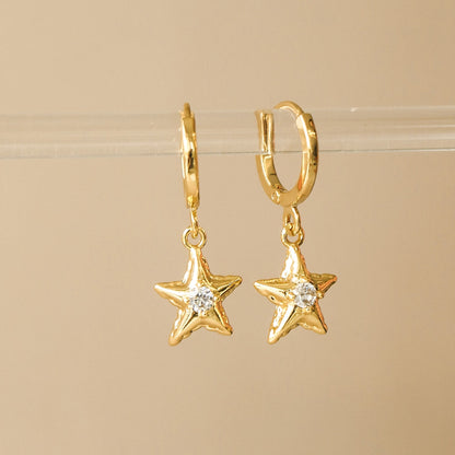 Anneau en laiton plaqué or avec un pendentif en forme d'étoile de mer avec un zircon en son centre. Les boucles d'oreilles sont accrochées sur un présentoir en acrylique transparent sur sur un fond beige. 