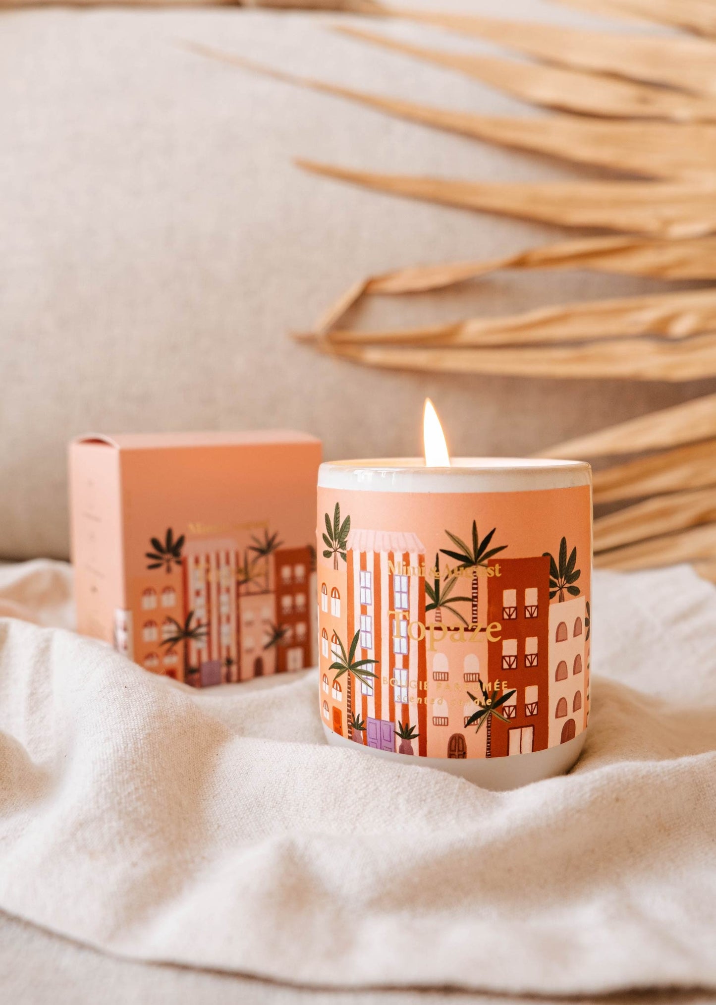 La bougie est coulé dans une tasse en céramique grise sans anse. Le fond de la tasse est arrondi. Sur la bougie, il y a une grosse étiquette sur laquelle on peut lire Mimi & August, Topaze, bougie parfumée, scented candle. Sur le fond de l'étiquette, on peut voir des grandes maison dans les tons brique, rose et orange. Il y plein de palmier autour. L'arrière-plan est un tissu beige et une feuille de palmier en papier. 