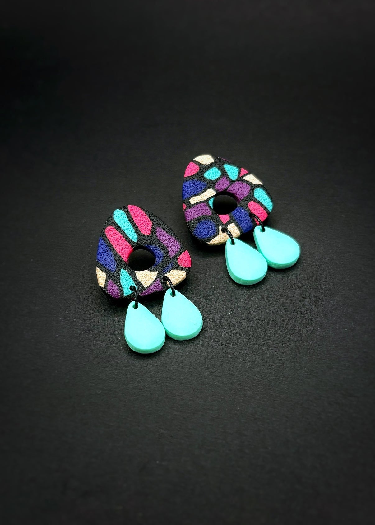 Petites boucles d'oreilles en triangle arrondi avec un trou au milieu. Les boucles d'oreilles sont noires, roses, mauves et bleus et font penser à un vitraille. Il y a deux petites pièces en forme de goute pendues en bas de chaque boucle d'oreille. L'arrière-plan est un fond noir. 