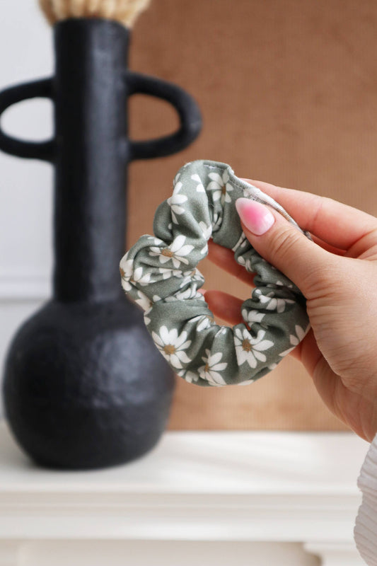 Chouchou en coton biologique Wild Daisy vert sauge tenu devant un vase noir, motif floral.