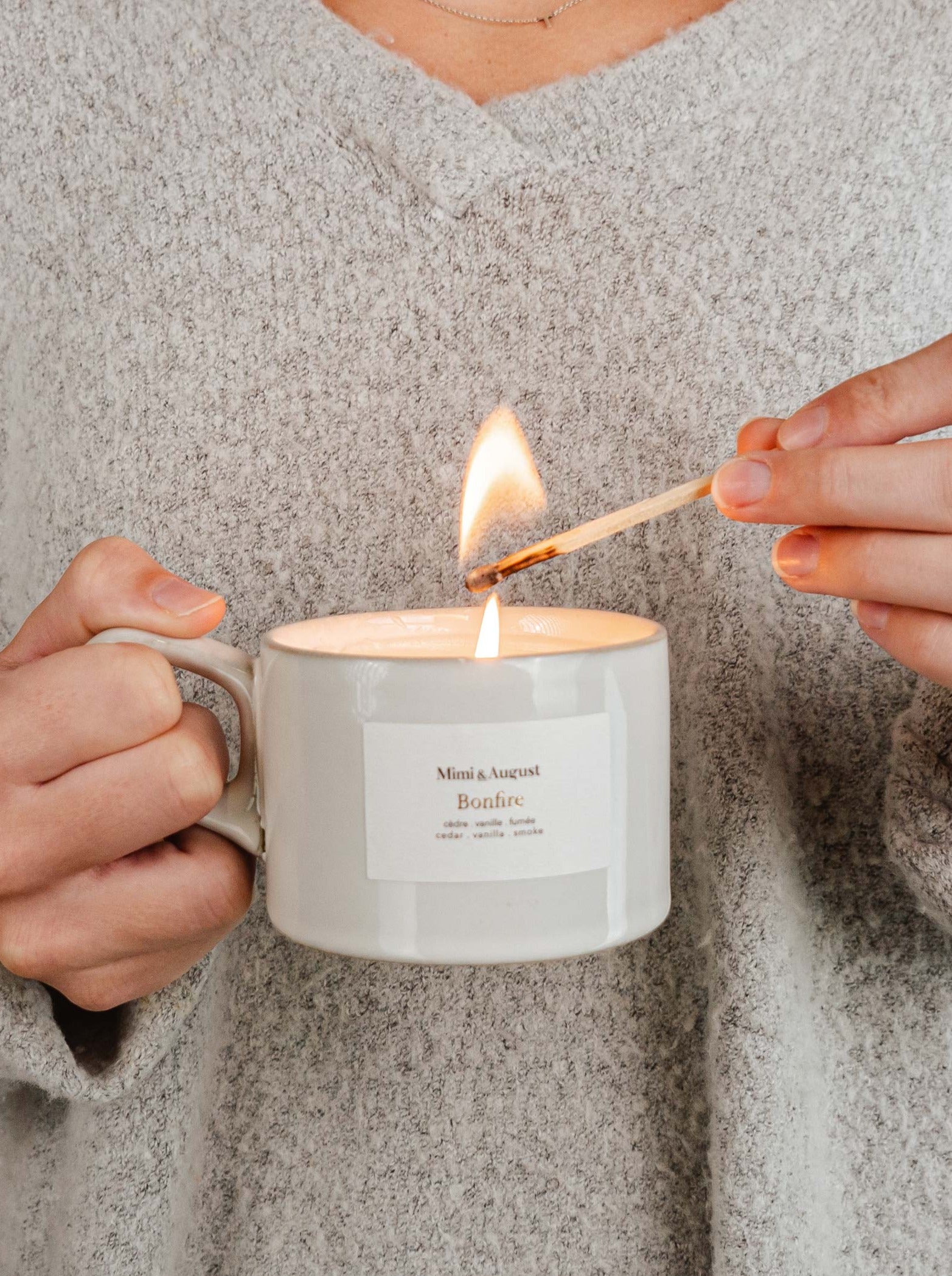 Tasse de taille moyenne en céramique beige avec une petite anse. Sur la tasse, il y a une étiquette blanche sur laquelle il est écrit Mimi & August, Bonfire et les notes de la bougie qui sont ici: cèdre, vanille et fumée. Sur la photo, on peut voir quelqu'un tenir la bougie d'une main et de l'autre l'allumer avec une allumette. La personne porte un pull gris. 